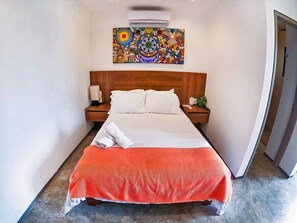 A hotel room with a bed, a nightstand, a wall-mounted air conditioner, and a colorful artwork above the bed. - Trendy Studio - Pool, Rooftop & Near Beach (Playa del Carmen)