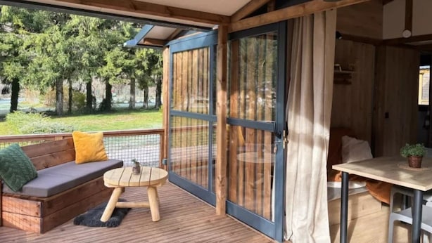 A wooden deck with a sofa, a small round table, and a dining area with a table and chairs. - ZEcamping 3* toboggans dog friendly locatif 36m2 5 pers (Chézery-Forens)