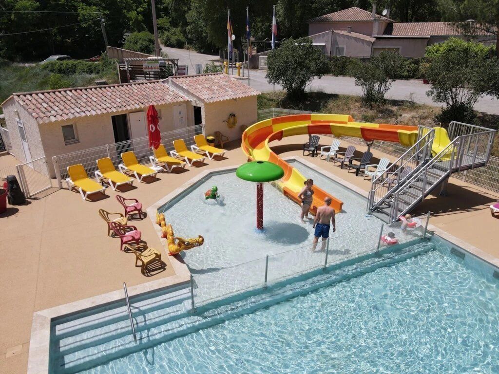 Uma piscina com escorrega, espreguiçadeiras e um pequeno edifício com guarda-sol vermelho.