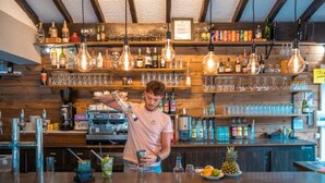 Un bar où un barman prépare un cocktail, des étagères remplies de bouteilles et de verres, et un panneau indiquant une réduction pour les paiements par carte bancaire.