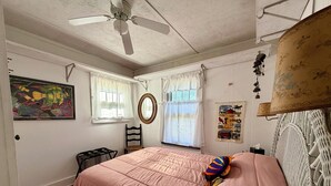 Un dormitorio con cama, ventilador de techo, ventana con cortinas, espejo montado en la pared y un cuadro enmarcado.