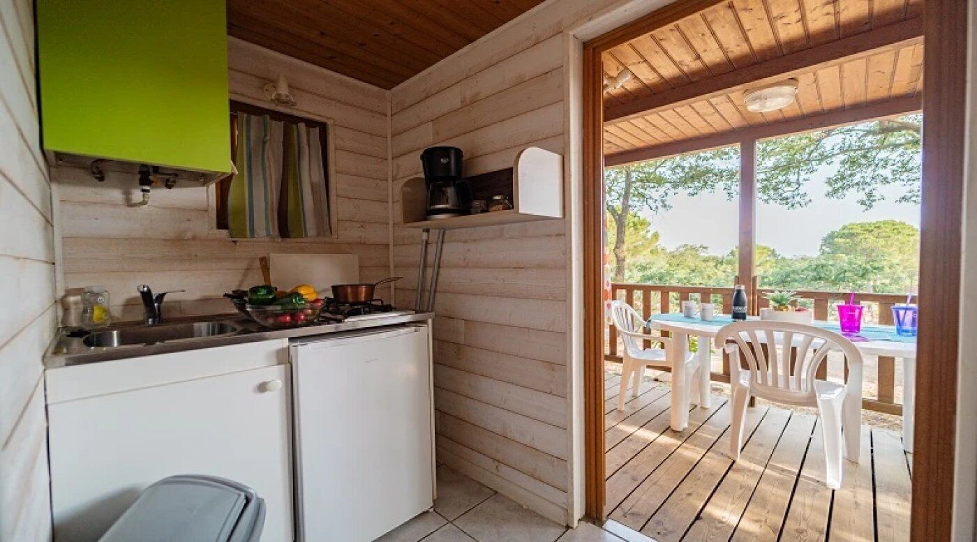 Une cuisine compacte équipée d’un réfrigérateur, d’un évier et d’une machine à café, le tout installé sur un plan de travail en bois. Une fenêtre est agrémentée de rideaux, et un espace extérieur aménagé offre des sièges pour profiter de l’extérieur.
