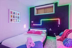 Une chambre moderne avec une tête de lit illuminée par des néons, un lit avec une literie rose et bleue, et une table de chevet avec des serviettes.
