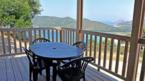 Una terraza de madera con una mesa redonda y cuatro sillas, con vista a un paisaje montañoso y una masa de agua.