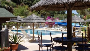 Un área de piscina con sillones de descanso y sombrillas de paja, rodeada de plantas en macetas y una terraza de madera.
