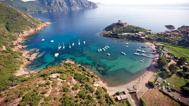 Vista aérea de una zona costera con aguas cristalinas, barcos y una pequeña isla con una estructura que parece un castillo.