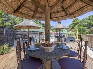 An outdoor dining area with wooden tables and chairs, thatched umbrellas, and a pool in the background.