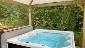 A hot tub in a covered outdoor area with a wooden deck and lush greenery in the background. - ZEcamping 3* piscine chauffée jacuzzi locatif TV 18m2 2 pers (Grand-Laviers)