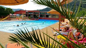 A swimming pool with people swimming and sunbathing, surrounded by palm trees and a building with a red roof. - ZEcamping 4* familial piscine pet friendly chalet TV BBQ clim 45m2 (Saint-Rome-de-Tarn)