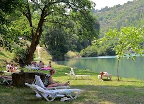 Un paesaggio con un lago, persone che si rilassano su sedie a prato e un piccolo albero.