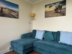 A teal sofa with multiple cushions, a lamp on a shelf, and two landscape paintings on the wall. - Chalet Riki Family Retreat in East Riding of Yorkshire (Withernsea)
