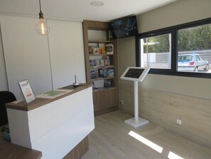 A reception desk with a computer, a bookshelf, a standing fan, and a window with a view of parked cars. - ZEcamping 4* animé 1km plage piscine couverte locatif TV 31m2 (Saint-Jean-de-Monts)