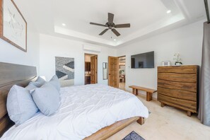 A bedroom with a bed, a ceiling fan, a wooden dresser, a TV, and a door leading to another room. - CCV Quivira - Luxurious and Beautiful 2BR w/ Ocean Views and Golf! (Cabo San Lucas)