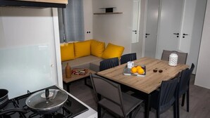 Ett kompakt kök med en spis, ett matbord med stolar, en soffa med gula kuddar och en liten hylla.