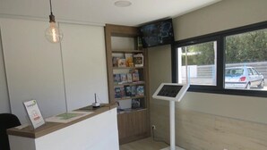 A reception desk with a computer, a bookshelf, a standing fan, and a window with a view of parked cars. - ZEcamping 4* 1km plage piscine couverte chiens acceptés mobil-home TV (Saint-Jean-de-Monts)