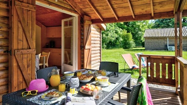 A wooden cabin with a covered patio area featuring a table set for breakfast, outdoor seating, and a view of the surrounding greenery. - ZEcamping 3* animé piscine chauffée locatif TV LV clim 35m2 (Hauterives)