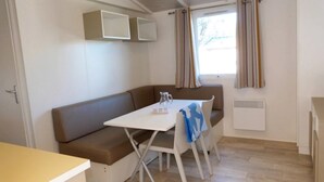 A compact living space with a sofa, a small table, and a window with curtains. - ZEcamping 3 étoiles 200m plage animaux acceptés locatif TV clim 26m2 (Hyères)