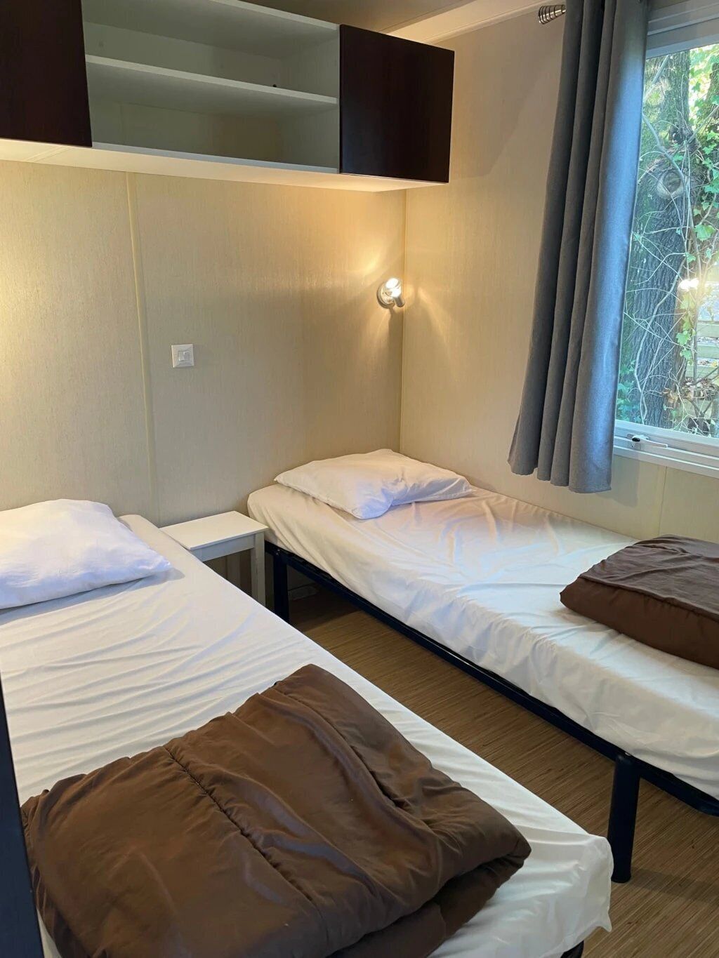 Ein Zimmer mit zwei Betten, einem an der Wand befestigten Regal, einem Nachttisch und einem Fenster mit Vorhängen.