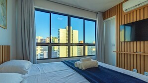 Een hotelkamer met een groot raam, een bed met witte lakens, een blauwe plaid en een flatscreen televisie aan de muur.