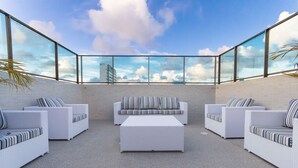 Een terras op het dak met witte rieten meubels en een glazen dak, met uitzicht op de lucht en wolken.