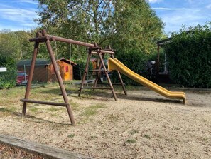 Une aire de jeux avec des balançoires et un toboggan, entourée d’arbres et d’une clôture.