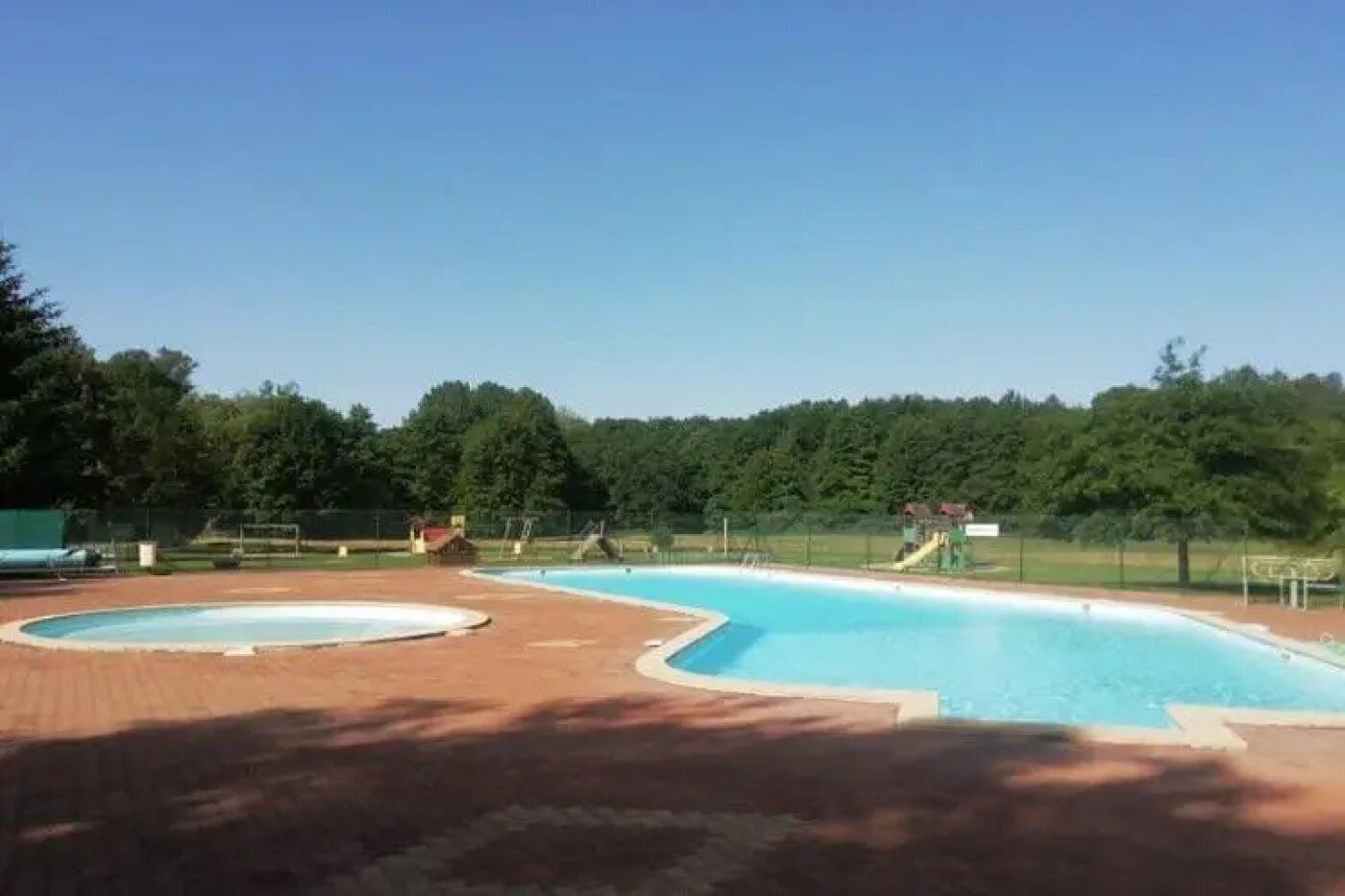 A swimming pool with two distinct sections, surrounded by a brick deck and a fenced area with trees in the background.