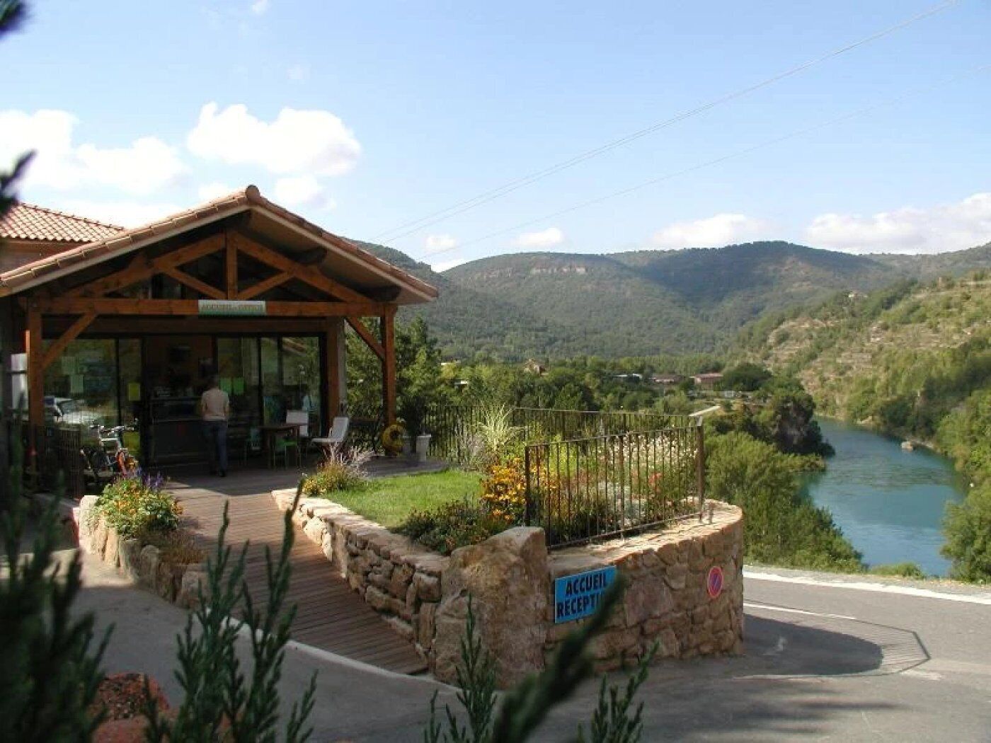 Edificio de recepción de madera con puerta de cristal, rodeado por un muro de piedra y una terraza de madera, con vistas a un río y montañas.