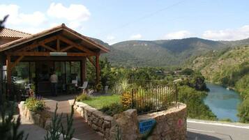 Edificio de recepción de madera con puerta de cristal, rodeado por un muro de piedra y una terraza de madera, con vistas a un río y montañas.