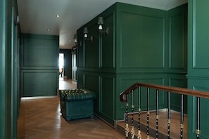 Un couloir aux murs recouverts de panneaux verts, un escalier en bois avec une rampe noire et un banc recouvert d’un tissu vert.
