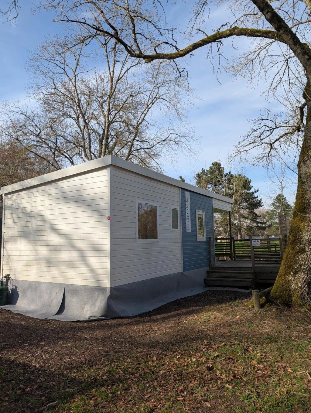 A small, single-story mobile home with a covered porch, situated in a wooded area with trees and a clear sky.