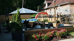 Een terras met houten tafels en stoelen, een overdekt zitgedeelte en een gebouw met een pannendak.