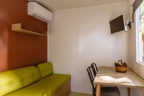 A small room with a green sofa, a wooden table, a television mounted on the wall, and a window. - ZEcamping 4* 3km plage piscine chauffée locatif TV clim 18m2 (Canet-en-Roussillon)