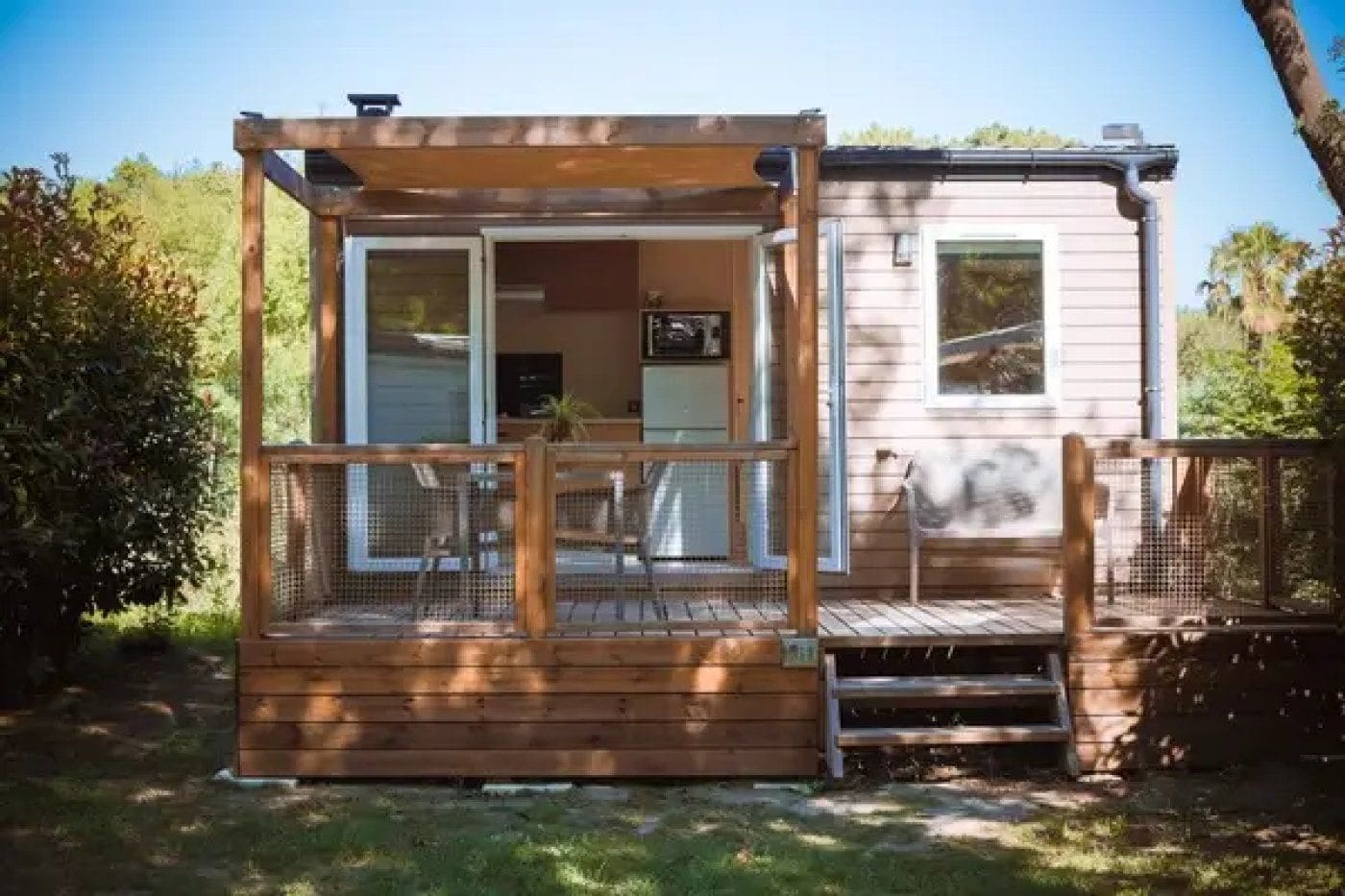 Una terraza de madera con zona de estar cubierta, un pequeño porche y un edificio de una sola planta con ventana y puerta.
