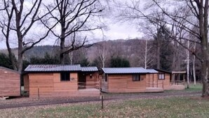 A wooden cabin with a porch and a small attached shed, surrounded by trees and a hilly landscape. - ZEcamping 3 étoiles piscine pet friendly club enfant tente 27m2 4 pers (Sauxillanges)