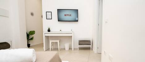 A modern, minimalist bedroom with a bed, a desk, and a TV mounted on the wall displaying 'NETFLIX'.
