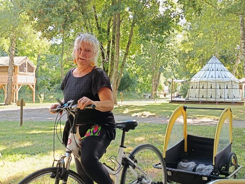A person riding a bicycle with a child seat attached, in a park with trees and wooden structures.