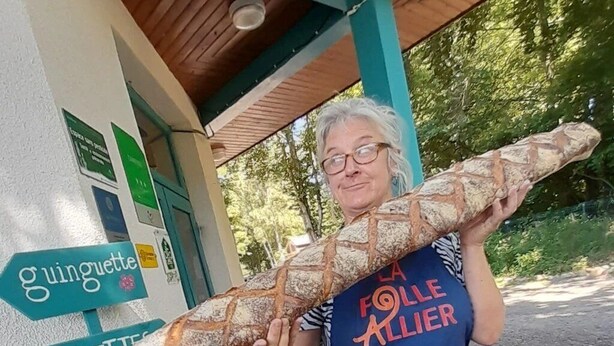 A person holding a large baguette in front of a building with signs indicating directions to 'gouguette' and 'toilettes'.