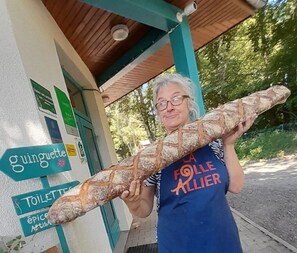A person holding a large baguette in front of a building with signs indicating directions to 'gouguette' and 'toilettes'. - ZEcamping 3 étoiles piscine animaux acceptés club enfant tente 2 pers (Sauxillanges)
