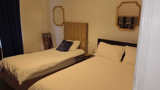 A hotel room with two beds, each with a wooden headboard and beige bedding. There is a bedside table with a lamp and a small decorative item on one bed. - PapaJoe's Get-away! Just what you deserve
sleeps 8/2 toilets/6 min to Waterworld (Stoke-on-Trent)