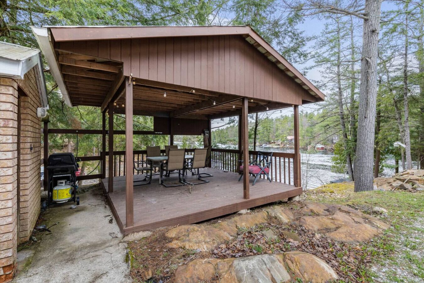 A covered outdoor deck with a table and chairs, a grill, and a view of a lake surrounded by trees.