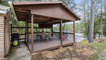 A covered outdoor deck with a table and chairs, a grill, and a view of a lake surrounded by trees.