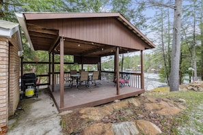 Une terrasse extérieure couverte, équipée d’une table et de chaises, d’un barbecue, et offrant une vue sur un lac entouré d’arbres.