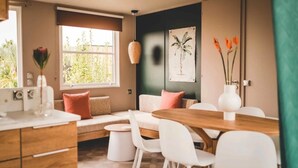 Ein modernes Wohnzimmer mit einem Holzesstisch, weißen Stühlen, einer Couch mit Kissen, einem großen Fenster und einer Wandpflanze.