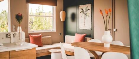 Un salon moderne avec une table à manger en bois, des chaises blanches, un canapé avec des coussins, une grande fenêtre et une plante fixée au mur.