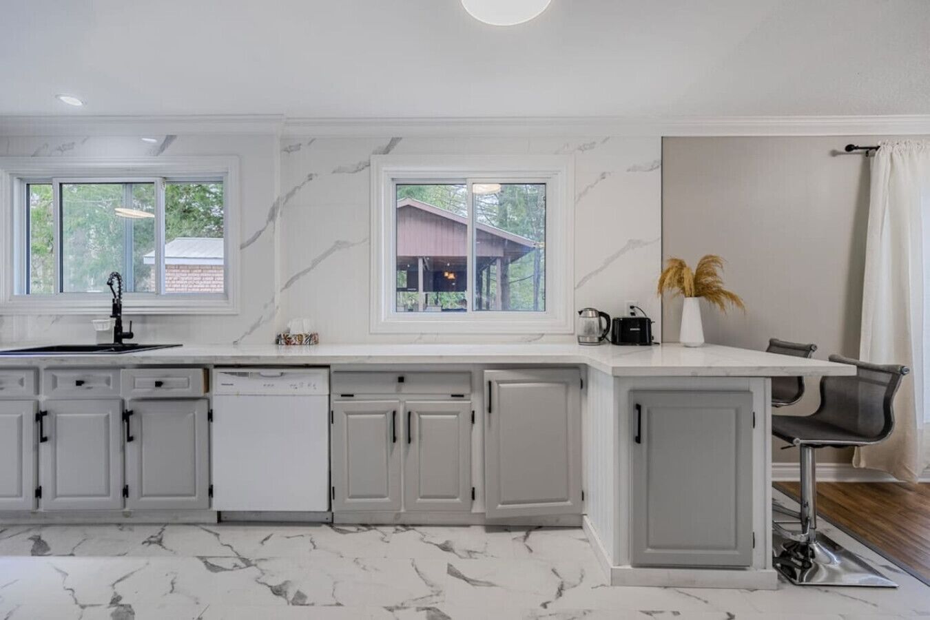 A modern kitchen with white cabinetry, a marble backsplash, and a large window overlooking a house.