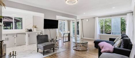 A spacious living area with a wooden floor, a grey sofa, a round glass coffee table, and a flat-screen TV mounted on the wall.