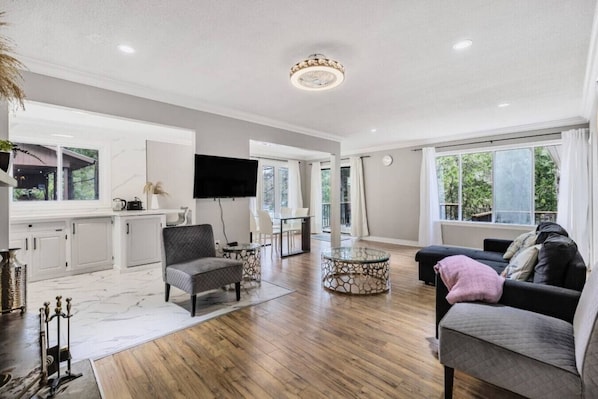 A spacious living area with a wooden floor, a grey sofa, a round glass coffee table, and a flat-screen TV mounted on the wall.