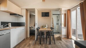 A compact kitchen with a dining area, a TV mounted above the table, and a sliding glass door leading to a balcony. - ZEcamping 4* espace aquatique animations locatif TV LV clim 32m2 (Soullans)