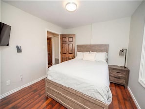 A bedroom with a bed, wooden headboard, bedside table, nightstand, and a wall-mounted TV.