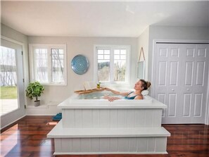 A person relaxing in a white hot tub in a room with large windows, a potted plant, and a door.